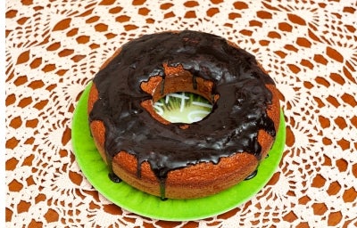 Bolo redondo recém-assado, com uma crosta dourada e cobertura de chocolate brilhante. Um pedaço foi cortado, revelando um interior amarelo úmido, e o bolo está sobre uma toalha de mesa de renda delicada.
