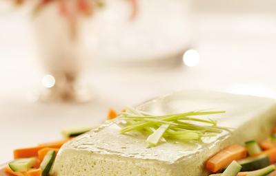 Fotografia em tons de branco, rosa e verde de uma bancada branca com uma bandeja rosa, sobre ela o terrine com palitos de cenoura e pepino. Ao fundo um vaso de flores rosa e uma jarra de água.