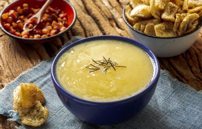 Fotografia de um recipiente fundo de vidro azul sobre um paninho azul claro apoiado em uma mesa de madeira. Dentro do recipiente tem caldo de aipim com um pouco de alecrim por cima, ao redor tem um pedaço de torrada caseira e amendoim.