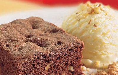 Fotografia em tons de marrom em uma bancada de madeira com uma foto aproximada de um prato polvilhado com cacau em pó, um brownie de chocolate e sorvete de creme.