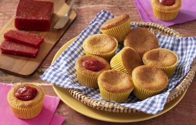 Fotografia em tons de marrom em uma bancada de madeira de cor marrom. No centro, um prato amarelo, uma cesta e um pano azul quadriculado em cima. Em cima do pano estão os cupcakes. Ao fundo, há uma tábua de madeira contendo fatias de goiabada e uma faca.