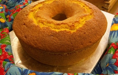 Fotografia de um bolo de fubá recém-assado, dourado e com textura uniforme, apresentado sobre uma superfície metálica. O bolo está cercado por um tecido colorido com padrão floral. Fotografia de um bolo de fubá recém-assado, dourado e com textura uniforme, apresentado sobre uma superfície metálica. O bolo está cercado por um tecido colorido com padrão floral.