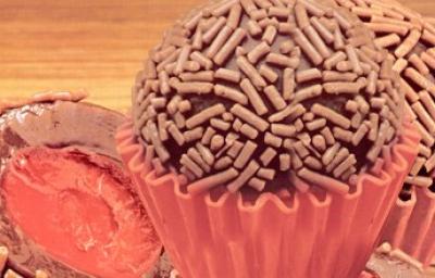 Fotografia em tons de marrom e vermelho de uma bancada de madeira com uma forminha vermelha, sobre ela brigadeiros recheados com cereja. Fotografia em tons de marrom e vermelho de uma bancada de madeira com uma forminha vermelha, sobre ela brigadeiros recheados com cereja.