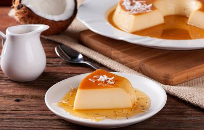 Fotografia de uma fatia de pudim de coco com caramelo salgado sobre um prato branco de vidro. Ao lado tem uma tábua de madeira com outro prato branco e o resto do pudim. Fotografia de uma fatia de pudim de coco com caramelo salgado sobre um prato branco de vidro. Ao lado tem uma tábua de madeira com outro prato branco e o resto do pudim.