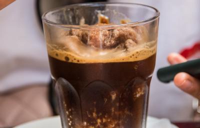Fotografia de uma granita feita de café, leite e gelo que está em um copo de vidro, o qual está apoiado sobre um miniprato de vidro branco. Ao fundo, tem alguém segurando uma colher com a sobremesa. Fotografia de uma granita feita de café, leite e gelo que está em um copo de vidro, o qual está apoiado sobre um miniprato de vidro branco. Ao fundo, tem alguém segurando uma colher com a sobremesa.