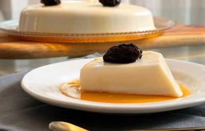 Fotografia mostra um pedaço de manjar de coco branco, com calda de caramelo e ameixa em cima.