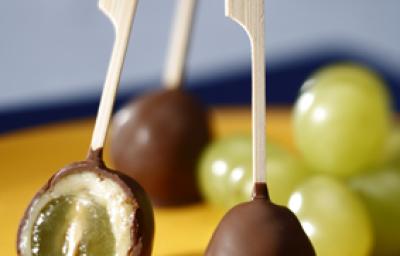 Fotografia em tons de amarelo, chocolate e azul, mostrando sobre bandeja amarela três espetinhos com chocolate, sendo um aberto e recheado com uva, algumas uvas verdes, tudo sobre guardanapo e bancada em tons de azul.