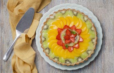 fotografia em tons de cinza, amarelo, verde e branco de uma bancada cinza vista de cima. Ao centro um prato redondo branco com uma torta com pedaços de frutas por cima para decorar, e ao lado um pano pardo cm uma espátula.