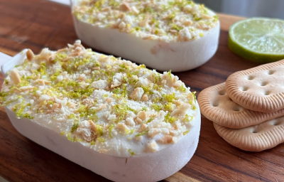 A imagem mostra dois sorvetes estilo picolé, claros, com uma textura crocante de biscoitos triturados e raspas de limão na superfície. Estão apoiados em uma tábua de madeira com 3 biscoitos de maisena e uma rodela de limão ao lado.