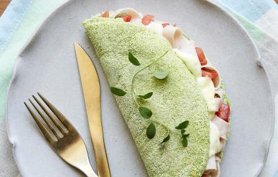 Fotografia em tons de verde em uma mesa de madeira com um pano listrado colorido em tons claro, um prato lilás redondo raso com uma tapioca verde de espinafre em cima dele. Fotografia em tons de verde em uma mesa de madeira com um pano listrado colorido em tons claro, um prato lilás redondo raso com uma tapioca verde de espinafre em cima dele.