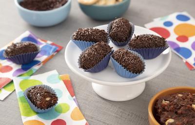 Foto da receita de Brigadeiro Croquete. Observa-se uma boleira pequena com 5 brigadeiros em formato de croquete em cima. A foto é decorada com guardanapos estampados de bolas coloridas e ao fundo observa-se dois potinhos: com brigadeiro e biscoito maria