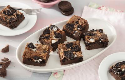 Foto da receita de Brownie de Leite Moça e Biscoito Negresco. Observa-se 7 fatias de brownie cortadas e dispostas em uma forma branca oval em um fundo rosa pastel.