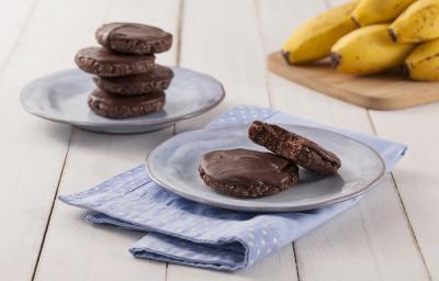 Fotografia em tons de marrom em uma bancada de madeira de cor branca. Ao centro, um pano azul claro estendido com um pires em cima contendo os cookies. Ao fundo, há mais um pires contendo cookies e uma tábua de madeira com algumas bananas. Fotografia em tons de marrom em uma bancada de madeira de cor branca. Ao centro, um pano azul claro estendido com um pires em cima contendo os cookies. Ao fundo, há mais um pires contendo cookies e uma tábua de madeira com algumas bananas.