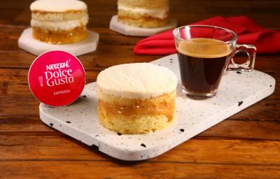 Fotografia em tons de vermelho em uma bancada de madeira escura, uma tábua de pedra branca com uma sobremesa de mini bolinho recheado com leite em pó e doce de leite. Ao lado, uma cápsula de Nescafé Dolce Gusto e uma xícara com o café pronto.