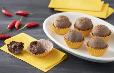 Fotografia em tons de amarelo de uma bancada cinza, sobre ela um prato oval com cinco trufas em forminhas amarelas, ao lado esquerdo um guardanapo amarelo com uma trufa cortada ao meio sobre ele. Ao fundo pimentas vermelhas. Fotografia em tons de amarelo de uma bancada cinza, sobre ela um prato oval com cinco trufas em forminhas amarelas, ao lado esquerdo um guardanapo amarelo com uma trufa cortada ao meio sobre ele. Ao fundo pimentas vermelhas.