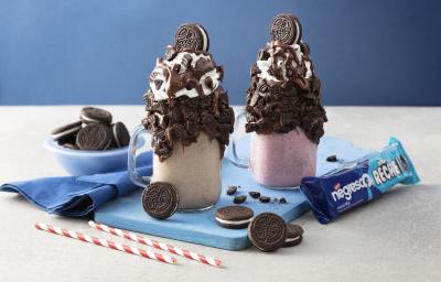 Fotografia em tons de azul, branco e marrom de uma bancada branca vista de frente, contém 2 copos transparentes um com milk-shake de baunilha e o outro de morango e ambos tem biscoitos de chocolates por cima para decorar. Ao lado o pacote do biscoito.