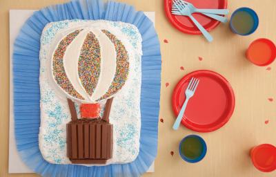 fotografia em tons de bege, vermelho e azul de uma bancada vista de cima. Contém uma tábua forrada de papel azul, com um bolo branco retangular com um balão por cima feito de creme e KITKAT. Ao lado pratos redondos vermelhos com garfos para servir.
