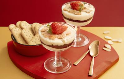 Foto da receita de Panacota de Frutas Vermelhas e Biscoito na taça. Observa-se duas taças com camadas e decoradas com lâminas de amêndoas e um morango por cima. Ao lado esquerdo, um potinho com biscoitos inteiros.
