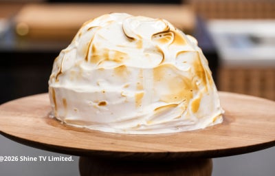 Sobremesa coberta com merengue tostado dourado sobre suporte de madeira, com fundo desfocado de cozinha.