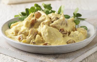 Fotografia em tons claros de uma salada de frango com molho ao curry e algumas folhas em um recipiente de cerâmica raso na cor branco. O prato está sobre uma peça de madeira em tom claro. Fotografia em tons claros de uma salada de frango com molho ao curry e algumas folhas em um recipiente de cerâmica raso na cor branco. O prato está sobre uma peça de madeira em tom claro.