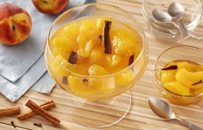 Fotografia em tons de amarelo em uma bancada de madeira de cor marrom. Ao centro, um recipiente de vidro contendo o doce de pêssego. Ao lado, há canelas em pau, um recipiente pequeno com doce e uma colher. Ao fundo, um pano azul claro com 2 pêssegos. Fotografia em tons de amarelo em uma bancada de madeira de cor marrom. Ao centro, um recipiente de vidro contendo o doce de pêssego. Ao lado, há canelas em pau, um recipiente pequeno com doce e uma colher. Ao fundo, um pano azul claro com 2 pêssegos.