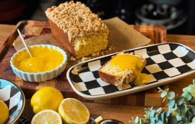 Fotografia de um bolo de limão com crumble apoiado em um papel manteiga, sobre uma tábua de madeira grande. Ao lado do bolo, assado em forma de bolo inglês, tem um prato preto e branco com uma faia do bolo e o curd. Tem um recipiente com lemon curd. Fotografia de um bolo de limão com crumble apoiado em um papel manteiga, sobre uma tábua de madeira grande. Ao lado do bolo, assado em forma de bolo inglês, tem um prato preto e branco com uma faia do bolo e o curd. Tem um recipiente com lemon curd.