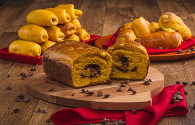 Foto de uma bancada de madeira. Sobre ela há uma tábua sobre um pano vermelho e nela está um pão em tom laranja aberto ao meio com recheio de chocolate. Ao fundo há mais pães menores e espalhadas pela bancada estão algumas gotas de chocolate Foto de uma bancada de madeira. Sobre ela há uma tábua sobre um pano vermelho e nela está um pão em tom laranja aberto ao meio com recheio de chocolate. Ao fundo há mais pães menores e espalhadas pela bancada estão algumas gotas de chocolate