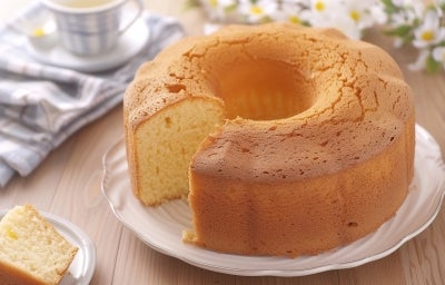 Um bolo redondo e dourado com furo no meio, servido sobre um prato branco com bordas. Uma fatia está cortada e colocada ao lado. O fundo tem uma xícara listrada, um guardanapo dobrado e flores brancas, criando um clima leve e acolhedor.