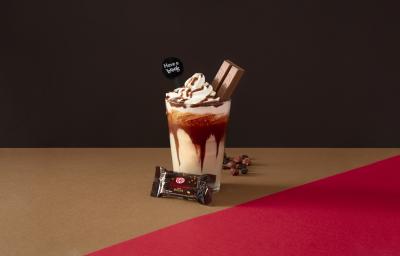 Foto da receita de Frapê Avelã. Observa-se Um fundo escuro marrom e ao centro um copo alto, decorado com calda de chocolate e dentro a bebida cremosa. Ao topo, chantilly e um KIT KAT Dark espetado. Avelãs decoram a parte direita do copo.