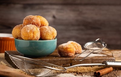 Fotografia de uma tigela de cerâmica teal cheia de bolinhas de massa frita, empilhadas de forma atraente. O cenário rústico inclui uma tábua de madeira polvilhada de açúcar e utensílios de cozinha.