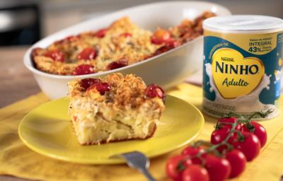 Fotografia em tons de amarelo em uma bancada de madeira, um pano amarelo e um prato amarelo redondo ao centro com um pedaço de torta de frango em cima. Ao fundo um recipiente com a torta inteira e a lata de Ninho Adulto ao lado. Fotografia em tons de amarelo em uma bancada de madeira, um pano amarelo e um prato amarelo redondo ao centro com um pedaço de torta de frango em cima. Ao fundo um recipiente com a torta inteira e a lata de Ninho Adulto ao lado.