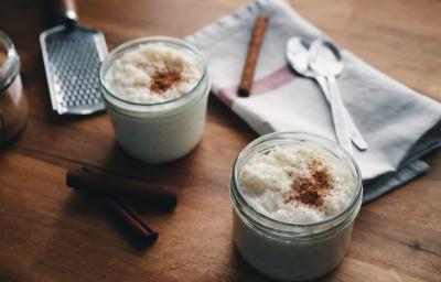 Fotografia de dois potes pequenos de vidro com arroz doce e canela polvilhada por cima. No fundo da foto, duas colheres de sopa e uma canela em pau sobre um pano de pano branco com uma listra vermelha. Ao lado dos potes, tem duas canelas em pau. Fotografia de dois potes pequenos de vidro com arroz doce e canela polvilhada por cima. No fundo da foto, duas colheres de sopa e uma canela em pau sobre um pano de pano branco com uma listra vermelha. Ao lado dos potes, tem duas canelas em pau.