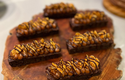 Fotografia em tons de marrom com uma bancada de madeira ao centro. Em cima da bancada existe 6 sobremesas com base de brownie e cobertas com um creme de caramelo e chocolate Fotografia em tons de marrom com uma bancada de madeira ao centro. Em cima da bancada existe 6 sobremesas com base de brownie e cobertas com um creme de caramelo e chocolate