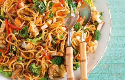 Foto de um prato branco cheio de yakissoba com frango e legumes. Encostado ao prato há duas colheres