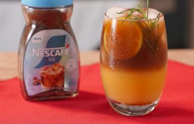 Fotografia de um copo de vidro com uma bebida de café gelado com tônica e laranja. A bebida está decorada com alecrim, e está ao lado de uma embalagem de nescafé gelado, sobre um pano vermelho. Fotografia de um copo de vidro com uma bebida de café gelado com tônica e laranja. A bebida está decorada com alecrim, e está ao lado de uma embalagem de nescafé gelado, sobre um pano vermelho.