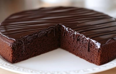 Em cima de uma mesa, prato redondo com bolo de chocolate redondo e cobertura de chocolate cremoso.