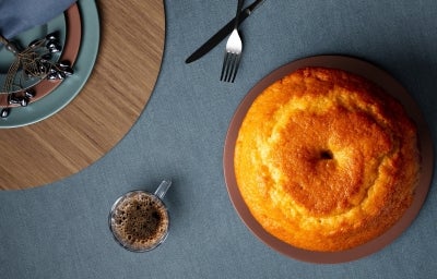 Fotografia de uma mesa elegantemente posta, com um bolo bundt dourado em um prato marrom. Ao lado, há pratos coloridos e um copo de café, com talheres dispostos de forma atraente. Fotografia de uma mesa elegantemente posta, com um bolo bundt dourado em um prato marrom. Ao lado, há pratos coloridos e um copo de café, com talheres dispostos de forma atraente.