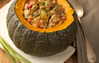 Fotografia em tons de marrom e cinza de uma bancada de madeira com um paninho branco, sobre ele uma abóbora recheada com carne e legumes. Ao lado cebolinhas e um paninho cinza com uma colher, ao fundo um prato branco. Fotografia em tons de marrom e cinza de uma bancada de madeira com um paninho branco, sobre ele uma abóbora recheada com carne e legumes. Ao lado cebolinhas e um paninho cinza com uma colher, ao fundo um prato branco.