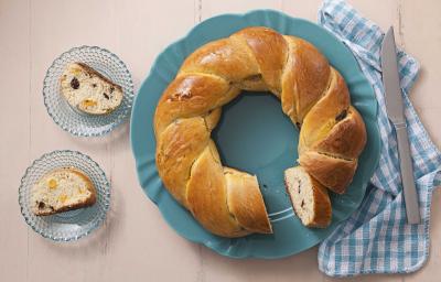 foto em tons de bege e azul de uma bancada bege vista de cima, contém um prato redondo azul com a rosca ao lado um pano xadrez azul e branco com uma faca. Do outro lado dois pratos azuis ambos com um pedaço da rosca com frutas secas.