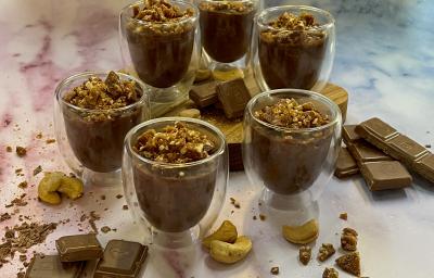 Foto da Receita de Mousse de Chocolate Crocante. Observa-se 6 taças de vidro individuais com a mousse dentro e com a pranilé de castanha de caju em cima decorando. Pedaços de chocolate e castanhas de caju decoram a foto também. Foto da Receita de Mousse de Chocolate Crocante. Observa-se 6 taças de vidro individuais com a mousse dentro e com a pranilé de castanha de caju em cima decorando. Pedaços de chocolate e castanhas de caju decoram a foto também.