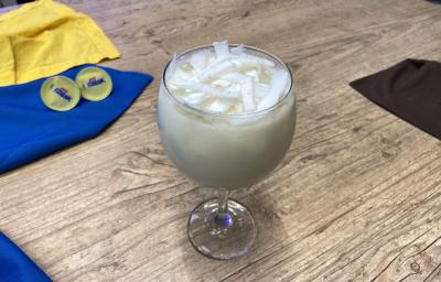Fotografia em tons de amarelo e azul em uma bancada de madeira, com paninhos amarelo e azul e uma taça de vidro ao centro com a bebida de coco e Galak dentro, decorado com lascas de coco fresco. Fotografia em tons de amarelo e azul em uma bancada de madeira, com paninhos amarelo e azul e uma taça de vidro ao centro com a bebida de coco e Galak dentro, decorado com lascas de coco fresco.