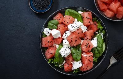 Fotografia vista de cima de uma salada feita com cubos de melancia, queijo, espinafre e gergelim. A salada está dentro de um recipiente fundo preto, sobre uma mesa preta.