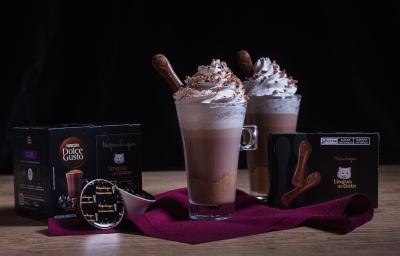 Fotografia em tons escuros de duas xícaras de vidro longas com Chococcino Mousse Língua de Gato, chantilly e língua de gato sobre um pano vinho. Ao lado tem uma caixa de nescafé e a outra de língua de gato. . Fotografia em tons escuros de duas xícaras de vidro longas com Chococcino Mousse Língua de Gato, chantilly e língua de gato sobre um pano vinho. Ao lado tem uma caixa de nescafé e a outra de língua de gato. .