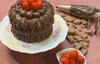 Foto em tons de marrom e vermelho da receita de bolo confeitado com ganache com morangos em cima, servido em um prato grande de porcelana branco com adornos em rosa. Ao lado, pedaços de chocolate e um bowl branco com morangos, além de um saco de confeitar