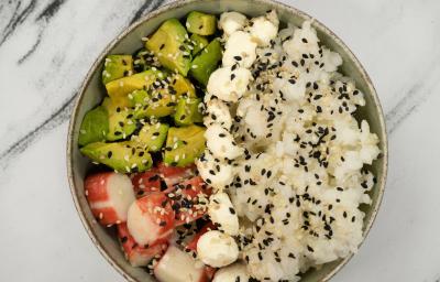 Fotografia em tons claros com um bowl ao centro. Dentro do bowl existe uma refeição a base de arroz e kani, temperada com gergelim e molho
