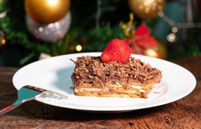 Fotografia de um pedaço de pavê de damasco com chocolate, cobertura de ganache, raspas de chocolate e um morango por cima. O pavê está em um prato raso branco, o qual está sobre uma mesa de madeira. Fotografia de um pedaço de pavê de damasco com chocolate, cobertura de ganache, raspas de chocolate e um morango por cima. O pavê está em um prato raso branco, o qual está sobre uma mesa de madeira.