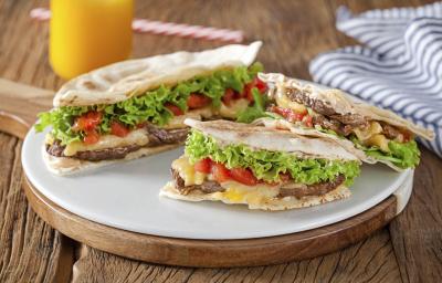 Fotografia em tons de branco e verde de bancada de madeira, tábua redonda com beirute partido ao meio, e uma das metades partidas ao meio mostrando a carne, queijo, tomate, alface e pão sírio. Ao fundo copo com suco laranja e pano listrado azul e branco. Fotografia em tons de branco e verde de bancada de madeira, tábua redonda com beirute partido ao meio, e uma das metades partidas ao meio mostrando a carne, queijo, tomate, alface e pão sírio. Ao fundo copo com suco laranja e pano listrado azul e branco.