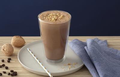 Foto em tons de marrom da receita de smoothie de café com nozes servida em um copo alto sobre uma base de porcelana cinza com um canudo listrado ao lado. Como decoração há duas nozes e um paninho azul