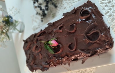 Fotografia do bolo de banana, chocolate e aveia assado em uma forma de bolo inglês, sobre um apoio retangular branco. O bolo tem uma cobertura de brigadeiro e decorado com raspas de chocolate.
