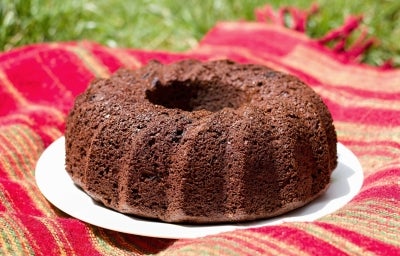 Fotografia de um bolo de chocolate em forma de bundt, com uma cor marrom profunda e textura densa, colocado em um prato branco. O fundo é um tecido listrado em vermelho e verde. Fotografia de um bolo de chocolate em forma de bundt, com uma cor marrom profunda e textura densa, colocado em um prato branco. O fundo é um tecido listrado em vermelho e verde.